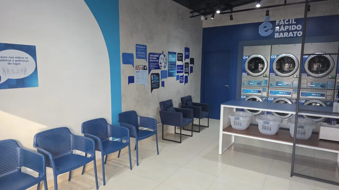 Vista ampla da sala da Leve & Lave: fileira de cadeiras, poltronas, mesa de dobrar com cestos e o mural de balões na parede de cimento.