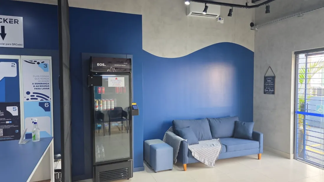 Sala de espera da Leve & Lave: sofá azul com manta, puff, geladeira de bebidas e ar-condicionado, na parede azul com a curva pintada.