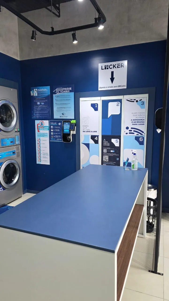 O Locker visto da bancada de dobrar da Leve & Lave, com as máquinas à esquerda e o totem de pagamento na parede azul.